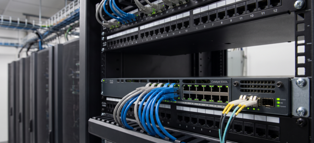 ภาพอุปกรณ์ Cisco Catalyst Switch ที่ติดตั้งอยู่ในตู้แร็คเซิร์ฟเวอร์ พร้อมเชื่อมต่อสายแลนสีฟ้าและไฟเบอร์ออปติกในห้องดาต้าเซ็นเตอร์